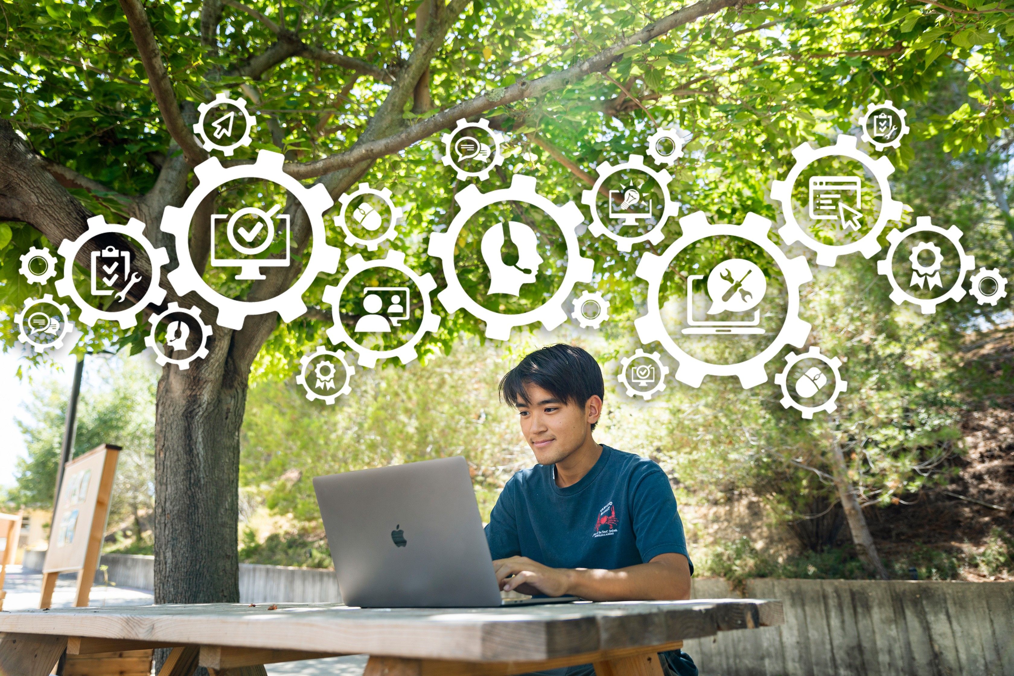 Cal Poly student sitting outdoors on a bench looking at their laptop screen. Random sized round gears with technology related icons floating above.